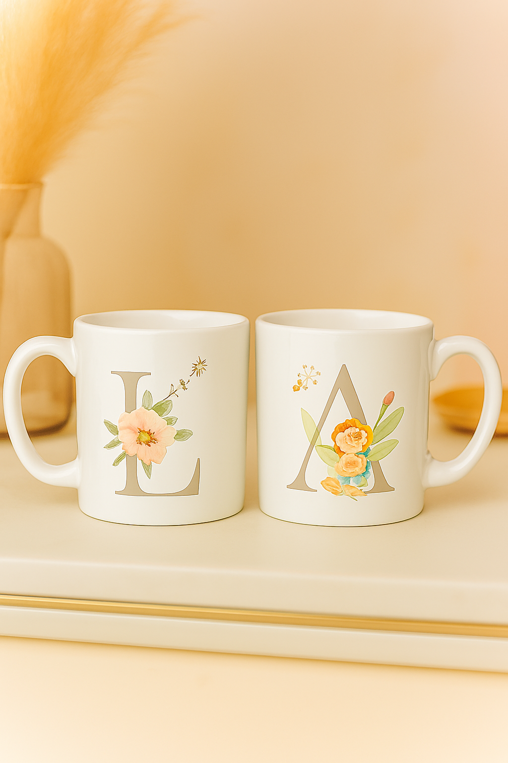 Two glossy white ceramic 11oz mugs with monogram letters “L” and “A,” featuring delicate floral and cherub illustrations. Set on a cream-toned surface with a soft beige and blush pink background, accented by pampas grass and a gold dish.