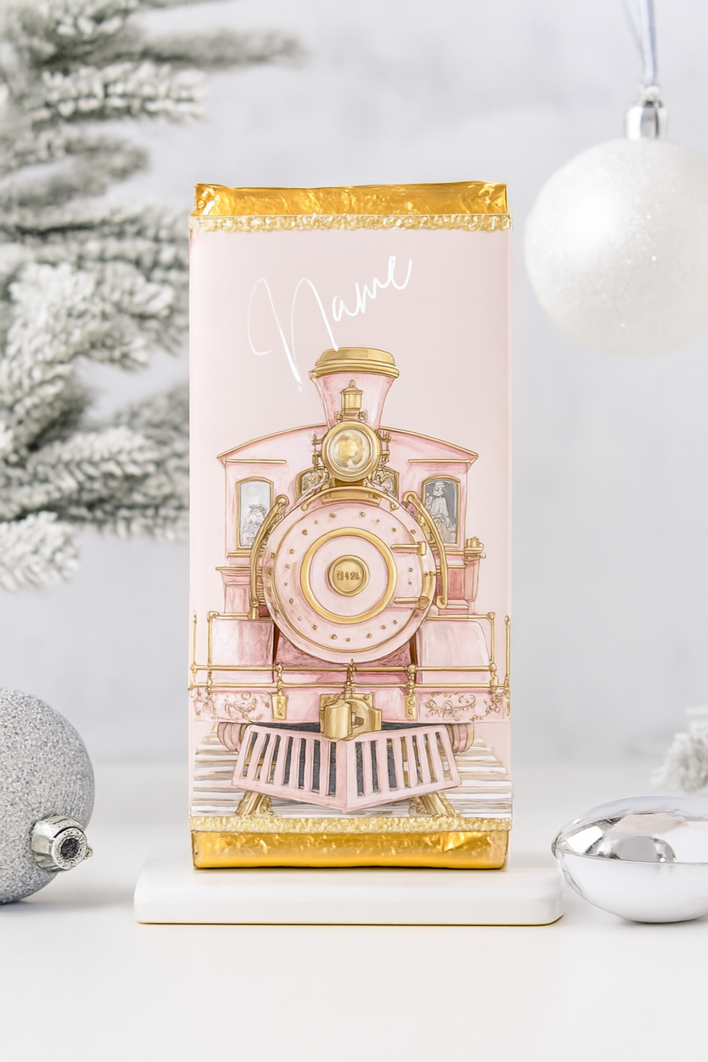 Personalised pink and gold Christmas train chocolate bar standing upright against a snowy white and silver festive background. Features a vintage steam locomotive illustration with ornate gold accents and a customisable name in white script.