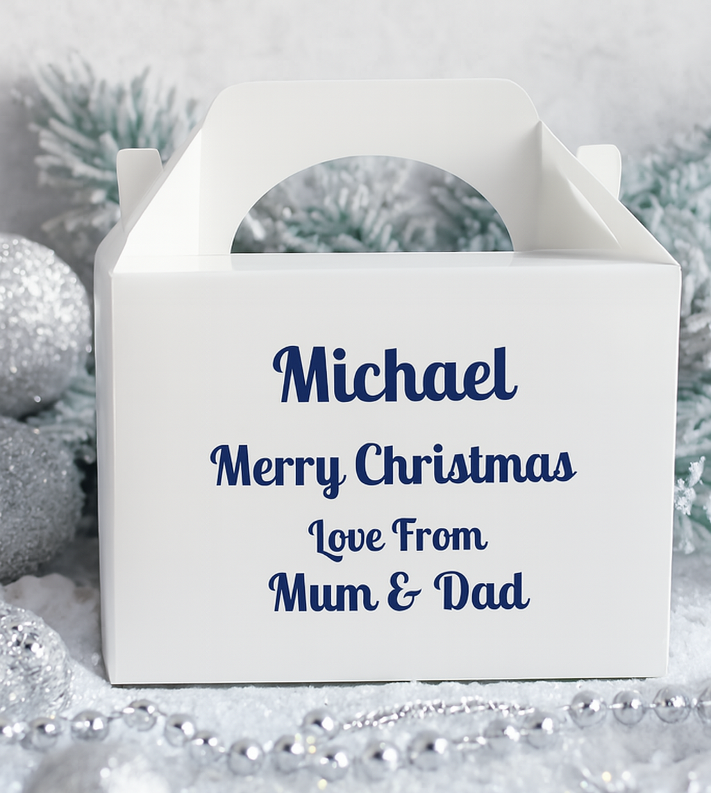 White personalised Christmas gift box with blue text reading “Michael Merry Christmas Love From Mum & Dad,” styled on a snowy silver background with glittery baubles, frosted pine branches, and silver bead garland for a festive, elegant presentation.