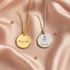 Two engraved pendant necklaces displayed on soft pink silk. The gold-plated pendant reads 'mum' in cursive, while the silver pendant features a flower and the word 'love'. Tiny round gold and silver balls accent the elegant scene.
