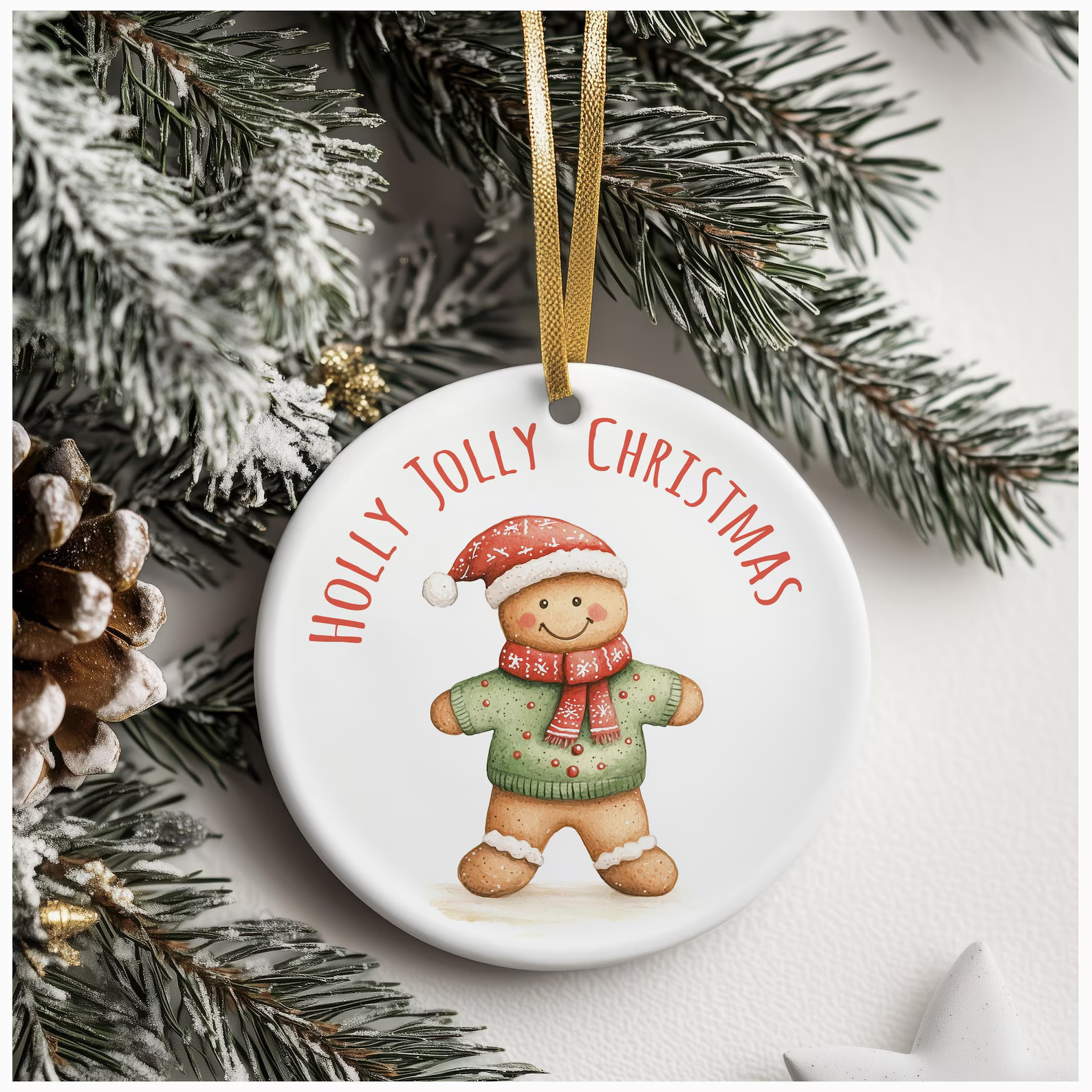 White ceramic ornament featuring a cheerful gingerbread figure in a green sweater and red scarf, with “HOLLY JOLLY CHRISTMAS” in red text, hanging from a gold ribbon on a snowy pine branch.