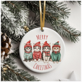 Round ceramic ornament with four cats dressed in winter sweaters and hats, surrounded by pine needles and a pine cone, with “MERRY CHRISTMAS” in red capital letters.