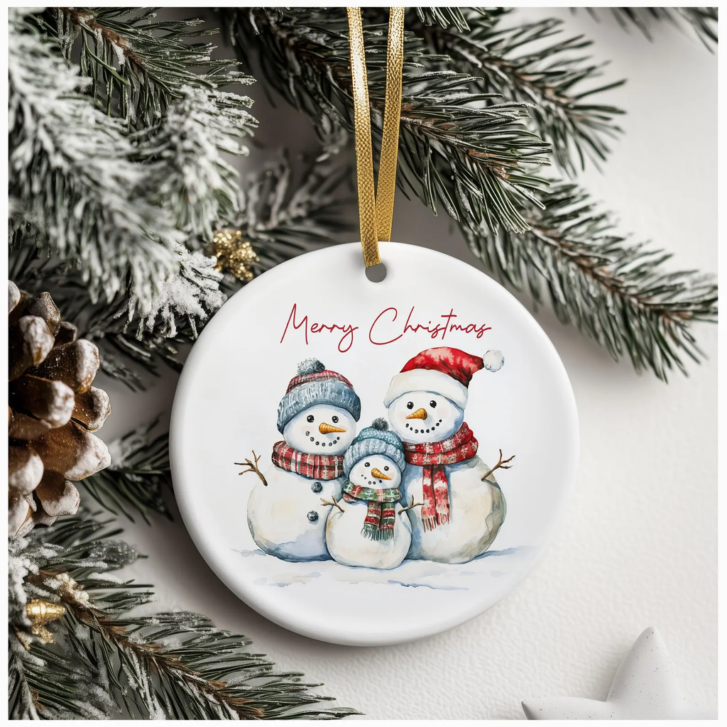 White ceramic Christmas ornament featuring three snowmen in festive hats and scarves, with “Merry Christmas” in red cursive, hanging from a gold ribbon on a snow-covered pine tree.