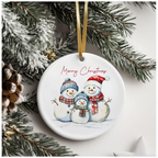 White ceramic Christmas ornament featuring three snowmen in festive hats and scarves, with “Merry Christmas” in red cursive, hanging from a gold ribbon on a snow-covered pine tree.