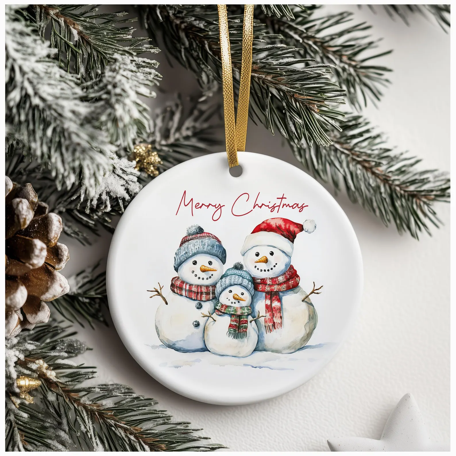 White ceramic Christmas ornament featuring three snowmen in festive hats and scarves, with “Merry Christmas” in red cursive, hanging from a gold ribbon on a snow-covered pine tree.